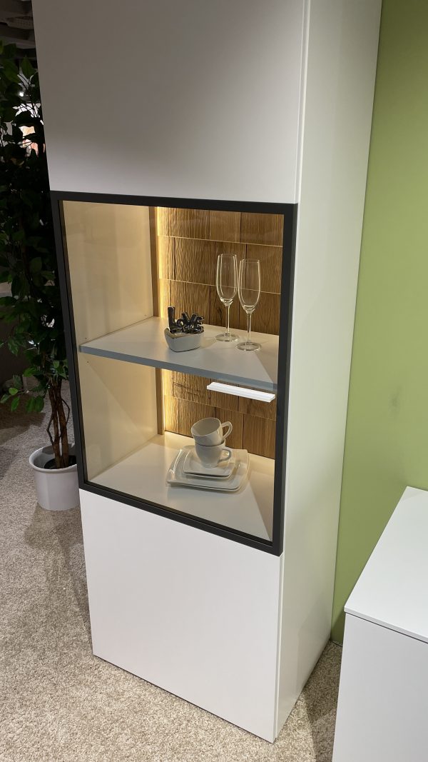 Wohnprogramm weiße Vitrine mit Glastüren hält Glaswaren, Teller und eine Tasse vor einer grünen Wand.