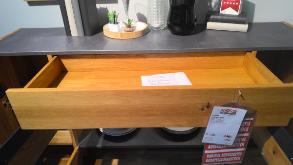 Der Schrank Anrichte Interliving-5603 hat eine offene Holzschublade, oben Dekor und eine Vase und unten Teller.