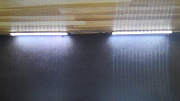 Zwei helle LED-Streifen unter der Anrichte Interliving-5603 werfen vertikale Lichtstreifen auf eine dunkle Wand.