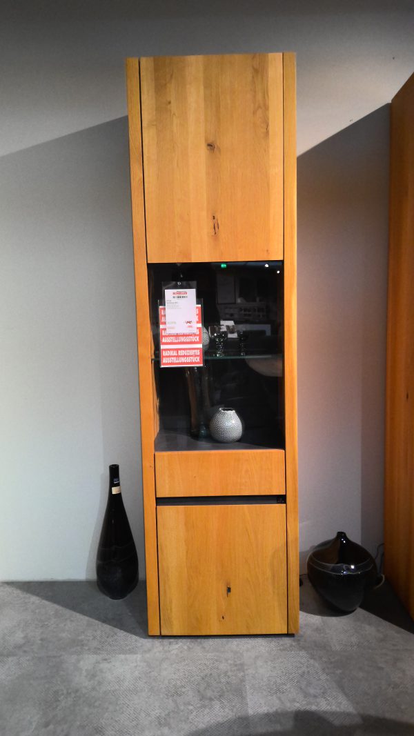 Anrichte Interliving-5603: Hohe Holzvitrine mit Glasvitrine, Vase darin und daneben platzierten Dekorationsartikeln.