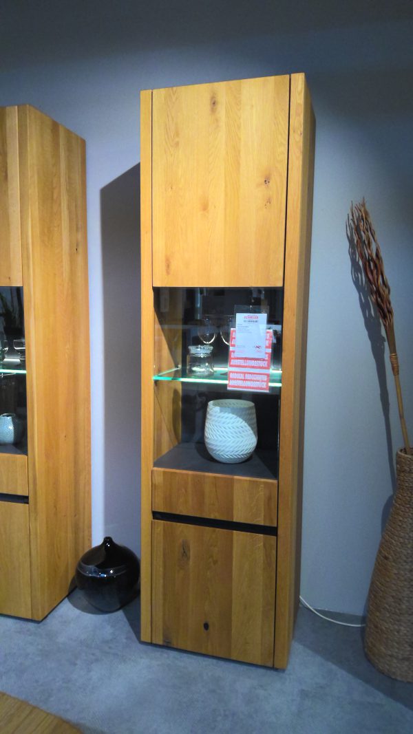Die Vitrine Interliving-5603 ist eine hohe Holzvitrine mit einem Glasboden, die mit einem weißen Dekorationsobjekt gezeigt wird.